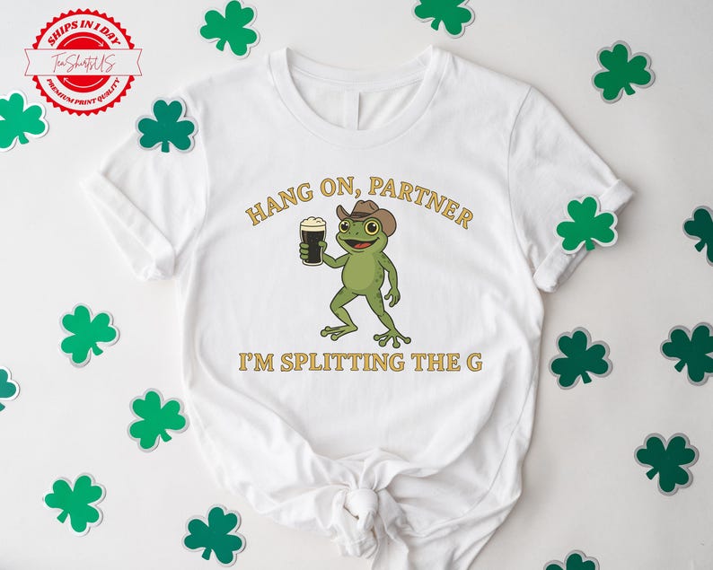 A white t-shirt with a cartoon frog holding a beer and the text "HANG ON PARTNER I'M SPLITTING THE G" printed on it. The t-shirt is surrounded by shamrocks on a white background.