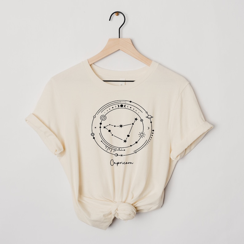 May include: A cream-colored t-shirt with a black graphic of the Capricorn zodiac sign surrounded by celestial symbols. The text "Capricorn" is printed below the graphic.