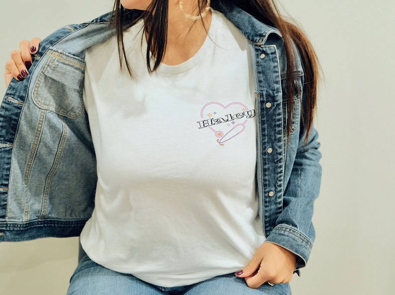 K&ouml;nnte beinhalten: Ein wei&szlig;es T-Shirt mit einem rosafarbenen Herzdesign, auf dem der Name "Haley" in schwarzer Schrift steht. Das Herz hat einen rosafarbenen Umriss und ein kleines, stilisiertes Skateboard im Inneren. Das T-Shirt wird von einer Person getragen, die eine blaue Jeansjacke tr&auml;gt.