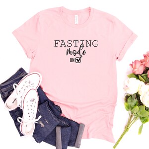 May include: A light pink t-shirt with the words "FASTING Mode ON" in black. The shirt is paired with blue jeans and white sneakers. A bouquet of pink and white flowers is also present.