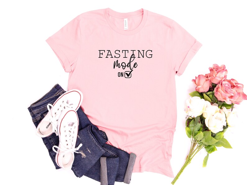May include: A light pink t-shirt with the words "FASTING Mode ON" in black. The shirt is paired with blue jeans and white sneakers. A bouquet of pink and white flowers is also present.