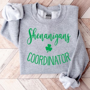 May include: A heather gray sweatshirt with the words "Shenanigans Coordinator" in green script, along with a green shamrock. The sleeves are folded, and the Gildan brand label is visible.