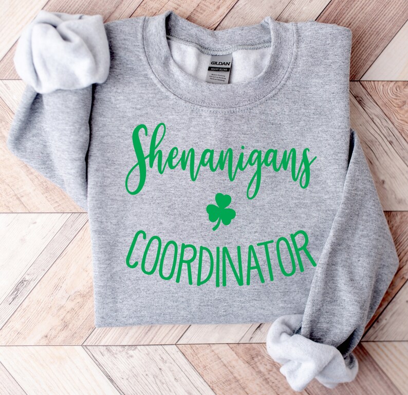 May include: A heather gray sweatshirt with the words "Shenanigans Coordinator" in green script, along with a green shamrock. The sleeves are folded, and the Gildan brand label is visible.