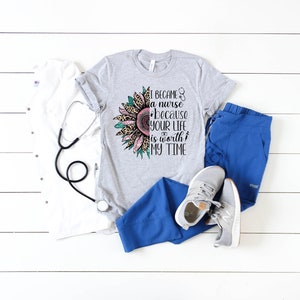 May include: A gray t-shirt with a sunflower graphic and the text "I became a nurse because your life is worth my time". The sunflower has pink and green petals with a leopard print center. The shirt is paired with blue scrub pants and white sneakers.