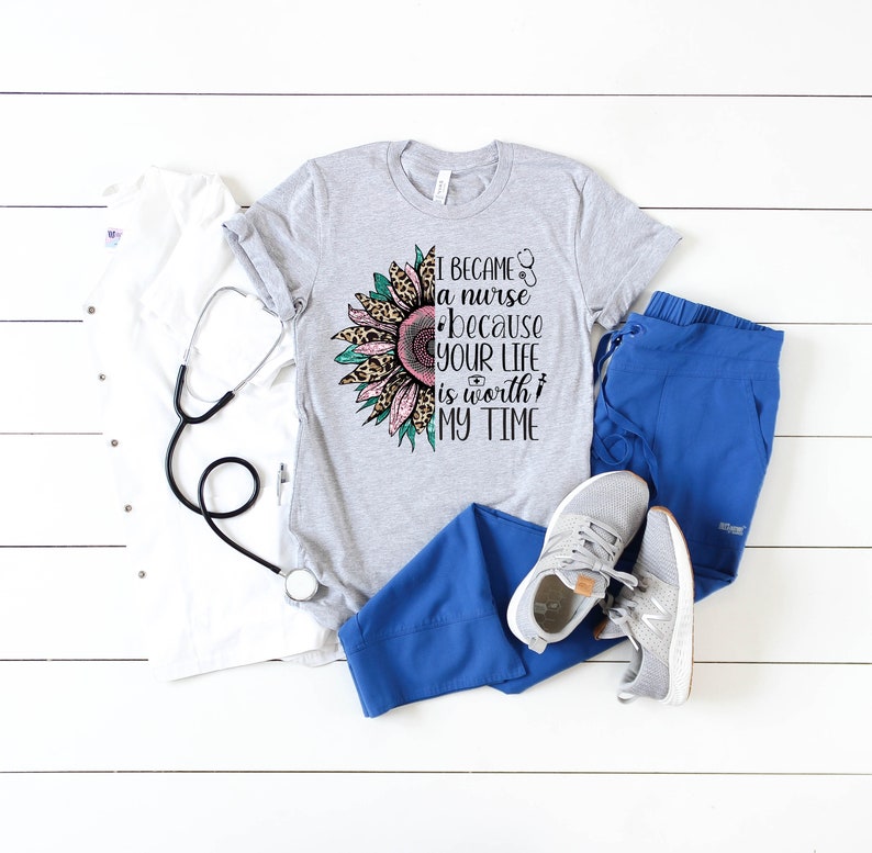 May include: A gray t-shirt with a sunflower graphic and the text "I became a nurse because your life is worth my time". The sunflower has pink and green petals with a leopard print center. The shirt is paired with blue scrub pants and white sneakers.
