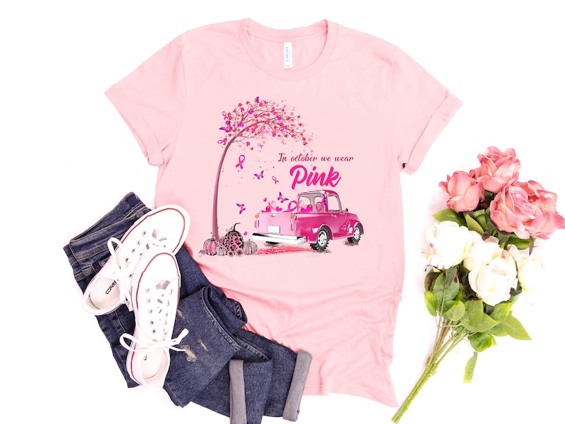 May include: Pink t-shirt with a pink truck carrying pumpkins and a pink ribbon tree. The text on the shirt reads "In October we wear Pink".