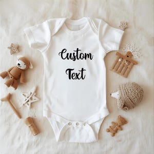 A white onesie with the text "Custom Text" printed on it, surrounded by various baby items such as a teddy bear, a star, and a hedgehog.