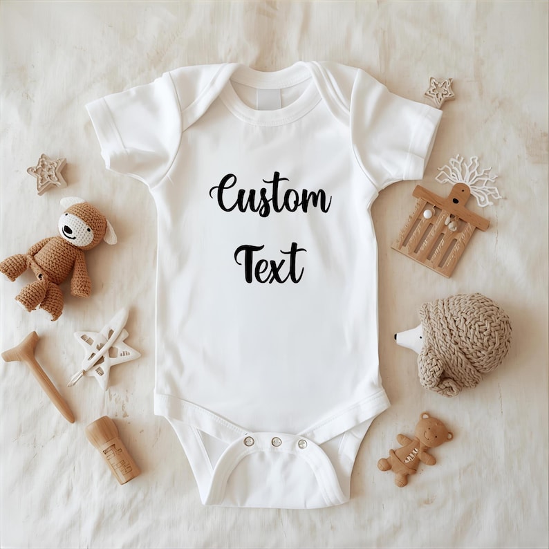 A white onesie with the text "Custom Text" printed on it, surrounded by various baby items such as a teddy bear, a star, and a hedgehog.