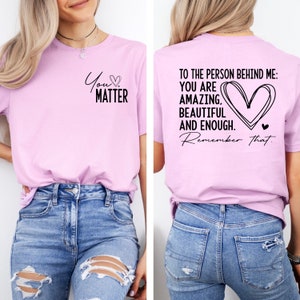 May include: A light pink t-shirt with the text "You Matter" and a heart on the front. The back of the shirt has the text "To the person behind me: You are amazing, beautiful and enough. Remember that." with a large heart design.