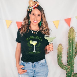 Puede incluir: Camiseta negra con un gr&aacute;fico de margarita y el texto "Margarita Social Club". La camiseta presenta una lima y una copa de c&oacute;ctel. La imagen tambi&eacute;n incluye un c&oacute;ctel de margarita en un vaso.