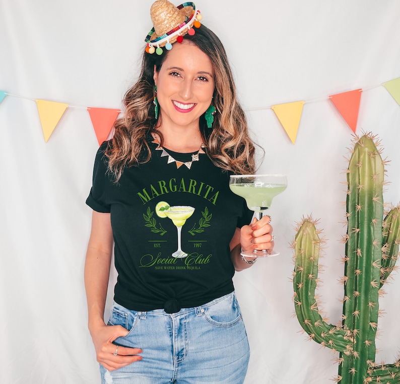 Puede incluir: Camiseta negra con un gr&aacute;fico de margarita y el texto "Margarita Social Club". La camiseta presenta una lima y una copa de c&oacute;ctel. La imagen tambi&eacute;n incluye un c&oacute;ctel de margarita en un vaso.
