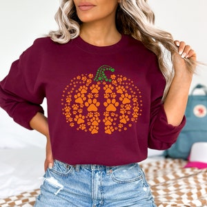 May include: A maroon sweatshirt featuring a pumpkin design made of orange paw prints. The pumpkin stem is green. The sweatshirt is a long-sleeved crew neck style.