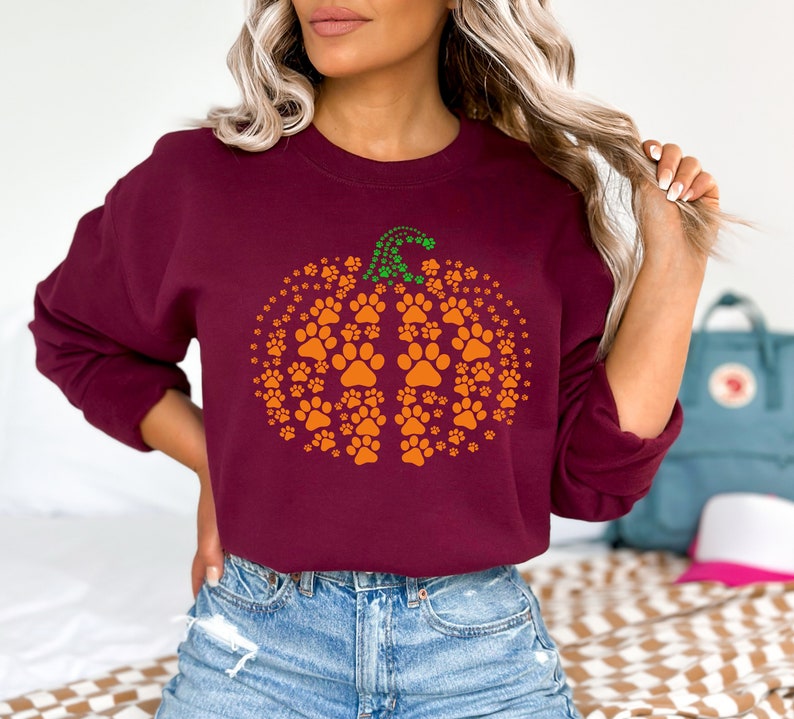 May include: A maroon sweatshirt featuring a pumpkin design made of orange paw prints. The pumpkin stem is green. The sweatshirt is a long-sleeved crew neck style.