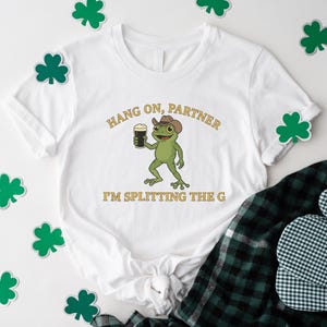 A white t-shirt with a cartoon frog wearing a cowboy hat and holding a beer, along with the text "HANG ON PARTNER I'M SPLITTING THE G" printed on it. The t-shirt is surrounded by shamrocks, and there is a plaid shirt and a pot holder visible in the background.