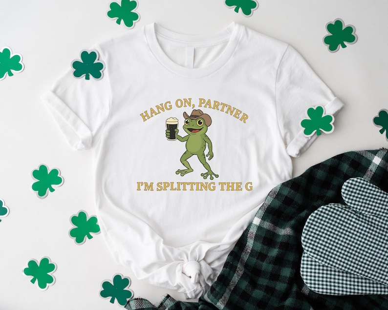 A white t-shirt with a cartoon frog wearing a cowboy hat and holding a beer, along with the text "HANG ON PARTNER I'M SPLITTING THE G" printed on it. The t-shirt is surrounded by shamrocks, and there is a plaid shirt and a pot holder visible in the background.