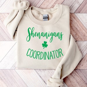 May include: A cream-colored sweatshirt with the words "Shenanigans Coordinator" in green script, along with a green shamrock. The sweatshirt is folded, with the sleeves rolled up. The Gildan brand tag is visible.