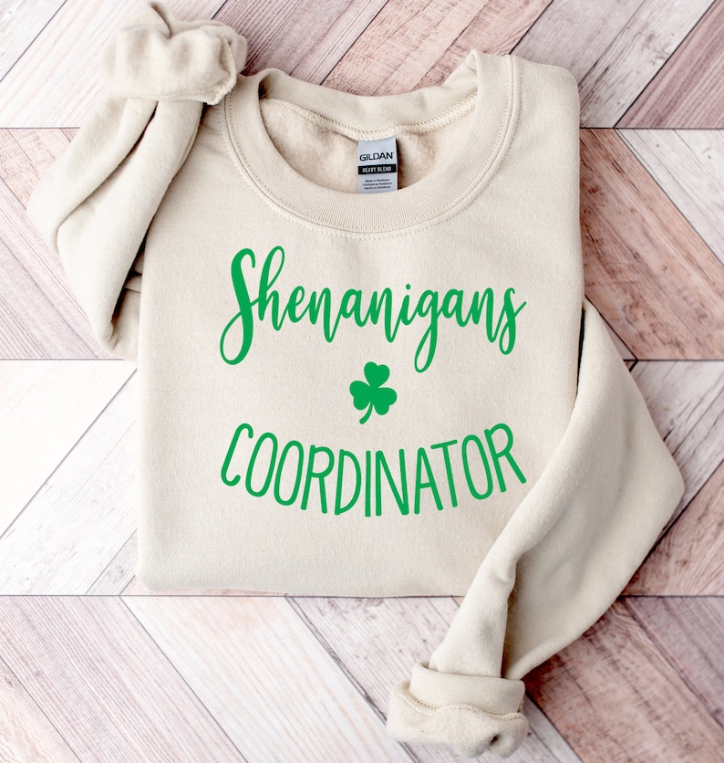 May include: A cream-colored sweatshirt with the words "Shenanigans Coordinator" in green script, along with a green shamrock. The sweatshirt is folded, with the sleeves rolled up. The Gildan brand tag is visible.