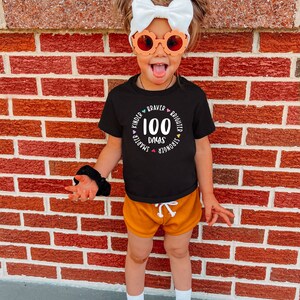 Op de afbeelding: Een kind draagt een zwart T-shirt met de tekst "100 dagen" in een cirkel met de woorden "Kinder, Smarter, Stronger, Braver, Brighter" eromheen. Het kind draagt een oranje broek en witte sneakers.