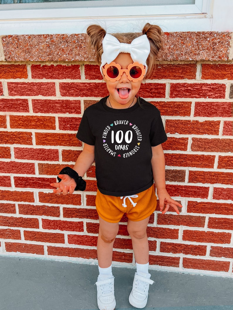 Op de afbeelding: Een kind draagt een zwart T-shirt met de tekst "100 dagen" in een cirkel met de woorden "Kinder, Smarter, Stronger, Braver, Brighter" eromheen. Het kind draagt een oranje broek en witte sneakers.