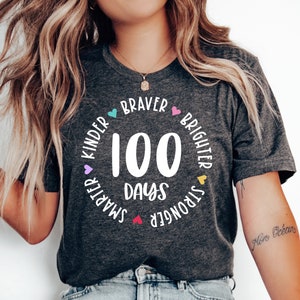 Op de afbeelding: Een grijs T-shirt met een wit cirkelvormig ontwerp met de tekst "100 dagen" en is omringd door de woorden "Kinder", "Braver", "Brighter", "Stronger" en "Smarter" met kleine hartjes tussen elk woord.