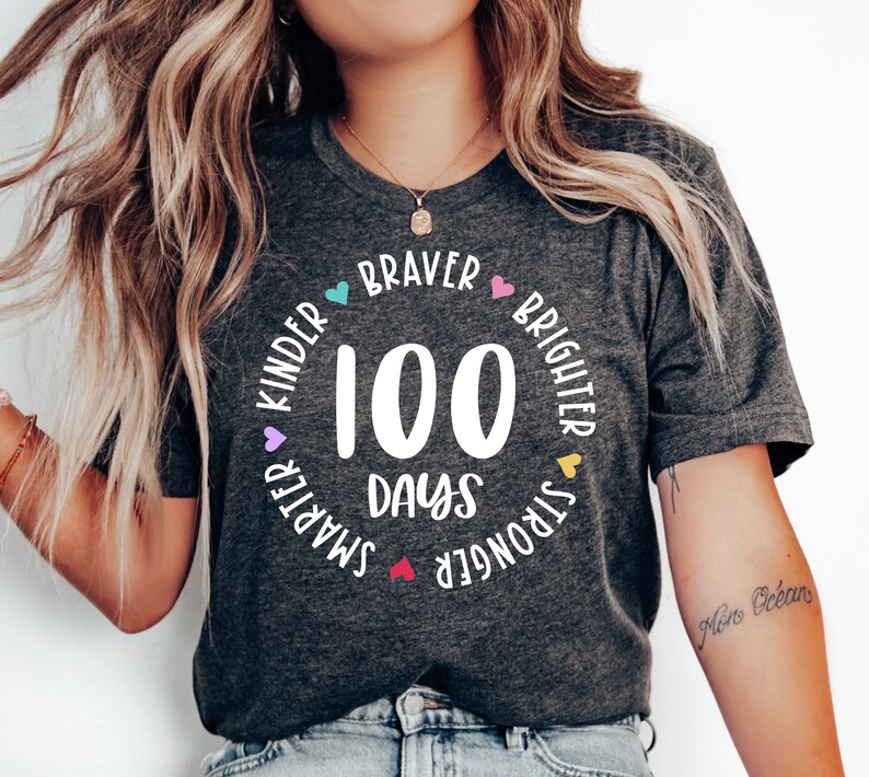 Op de afbeelding: Een grijs T-shirt met een wit cirkelvormig ontwerp met de tekst "100 dagen" en is omringd door de woorden "Kinder", "Braver", "Brighter", "Stronger" en "Smarter" met kleine hartjes tussen elk woord.