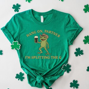 A green t-shirt with a cartoon frog and the text "HANG ON PARTNER I'M SPLITTING THE G" printed on it, along with shamrocks scattered around the shirt.