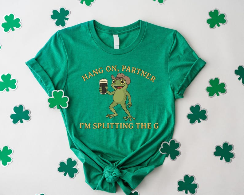 A green t-shirt with a cartoon frog and the text "HANG ON PARTNER I'M SPLITTING THE G" printed on it, along with shamrocks scattered around the shirt.