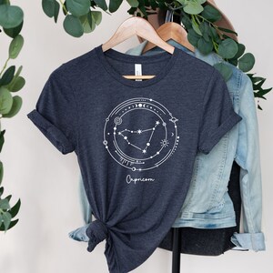 May include: A dark heather grey t-shirt with a white graphic of the Capricorn constellation surrounded by celestial symbols. The text "Capricorn" is printed below the graphic.