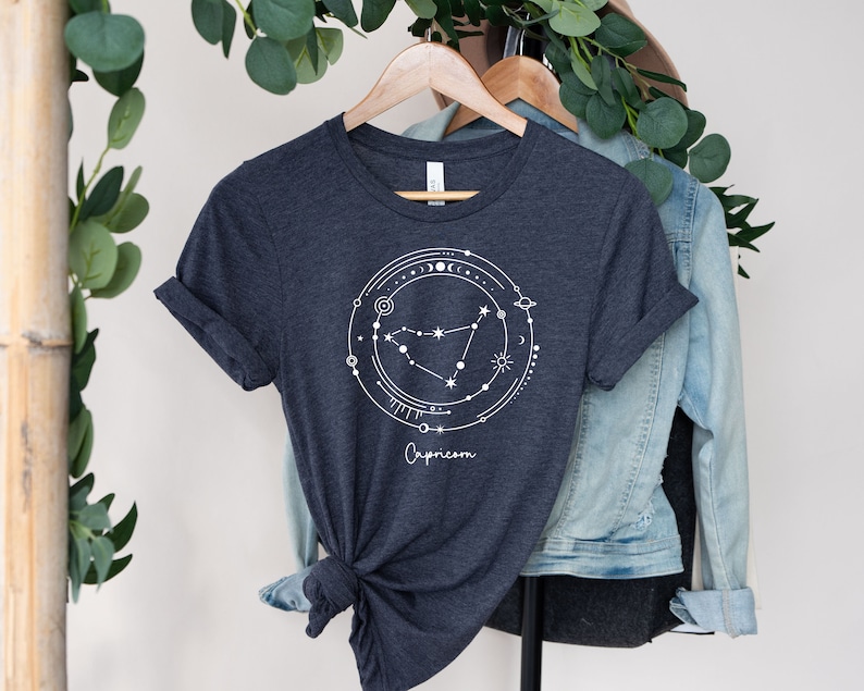May include: A dark heather grey t-shirt with a white graphic of the Capricorn constellation surrounded by celestial symbols. The text "Capricorn" is printed below the graphic.