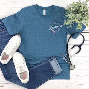 K&ouml;nnte beinhalten: Ein blaues T-Shirt mit einem rosafarbenen Herz und einem Stethoskop-Design. Der Text "Haley" befindet sich im Herzen. Das Shirt liegt auf einer wei&szlig;en Holzoberfl&auml;che mit einem Paar wei&szlig;en Sneakers und einer blauen Jeans.