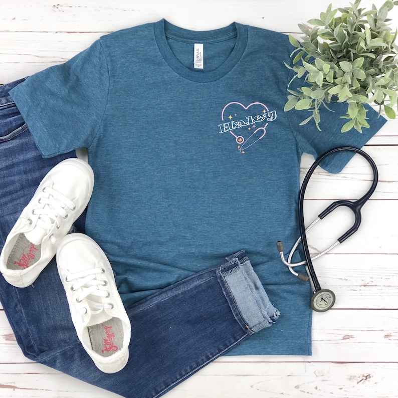 K&ouml;nnte beinhalten: Ein blaues T-Shirt mit einem rosafarbenen Herz und einem Stethoskop-Design. Der Text "Haley" befindet sich im Herzen. Das Shirt liegt auf einer wei&szlig;en Holzoberfl&auml;che mit einem Paar wei&szlig;en Sneakers und einer blauen Jeans.