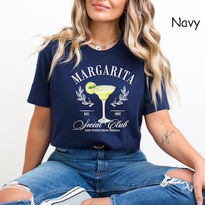Puede incluir: Camiseta azul marino con un dise&ntilde;o de margarita. El dise&ntilde;o incluye la palabra "Margarita" sobre una ilustraci&oacute;n de c&oacute;ctel, con el texto "Social Club" y "Save Water Drink Tequila". La camiseta tiene un cuello redondo cl&aacute;sico.