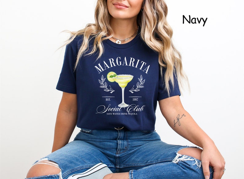 Puede incluir: Camiseta azul marino con un dise&ntilde;o de margarita. El dise&ntilde;o incluye la palabra "Margarita" sobre una ilustraci&oacute;n de c&oacute;ctel, con el texto "Social Club" y "Save Water Drink Tequila". La camiseta tiene un cuello redondo cl&aacute;sico.
