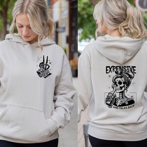 May include: A light beige hoodie with a black skeleton graphic on the back. The graphic shows a skeleton with a bandana and a drink in its hand. The text "Expensive and Talks Back" is below the graphic. The front of the hoodie has a black graphic of a hand with roses.