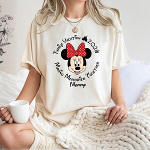 A woman wearing a white t-shirt with a Minnie Mouse design and text, sitting on a bed with a white blanket.