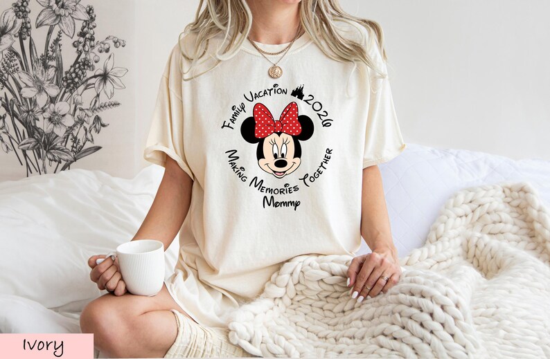 A woman wearing a white t-shirt with a Minnie Mouse design and text, sitting on a bed with a white blanket.