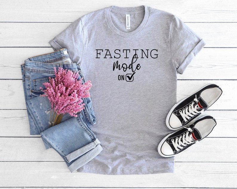 May include: A heather gray t-shirt with the text "FASTING mode ON" in black. The shirt is styled with blue jeans, pink flowers, and black canvas sneakers with white laces, all arranged on a white wooden surface.