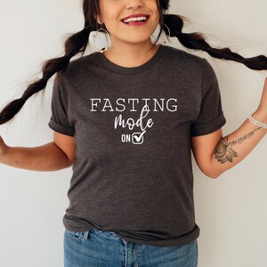 May include: Dark gray t-shirt with the text "FASTING mode ON" in white. The shirt has a rolled sleeve design. The text includes a checkmark symbol. The shirt is worn by a person with long dark braids.