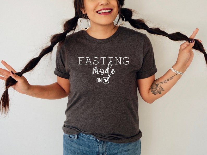 May include: Dark gray t-shirt with the text "FASTING mode ON" in white. The shirt has a rolled sleeve design. The text includes a checkmark symbol. The shirt is worn by a person with long dark braids.