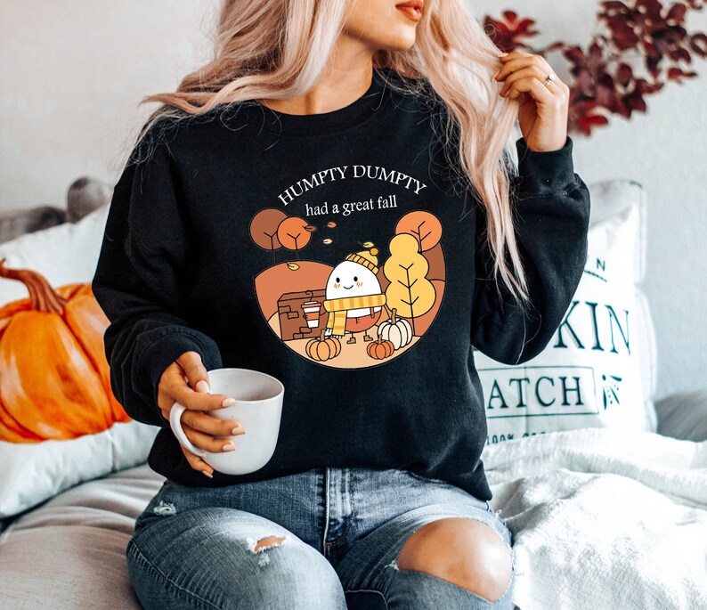 May include: A black sweatshirt with a white graphic of a cartoon egg wearing a scarf and hat, surrounded by fall leaves. The text "Humpty Dumpty had a great fall" is printed above the egg.