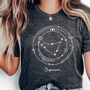 May include: A dark gray t-shirt with a white graphic of the Capricorn constellation surrounded by celestial symbols. The text "Capricorn" is printed below the graphic.
