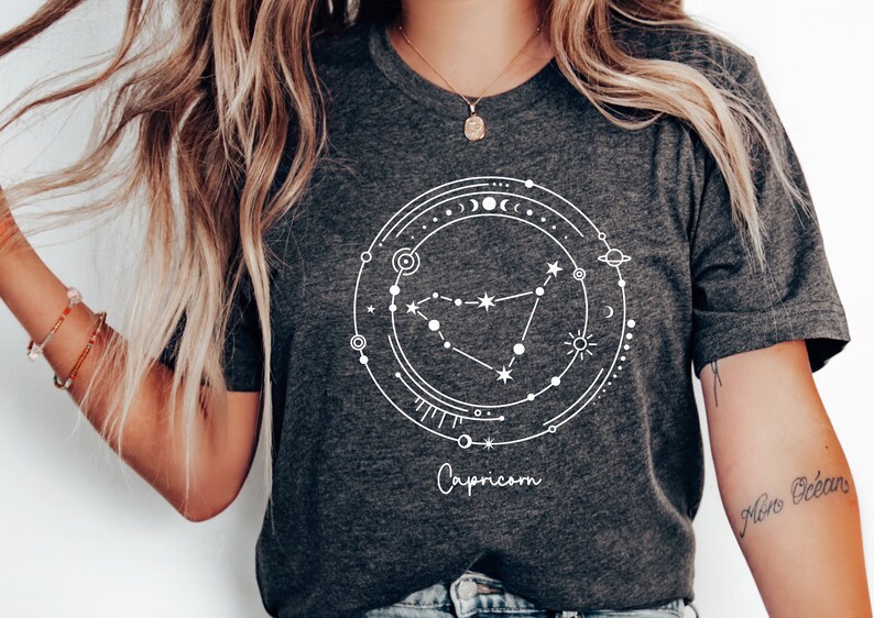 May include: A dark gray t-shirt with a white graphic of the Capricorn constellation surrounded by celestial symbols. The text "Capricorn" is printed below the graphic.