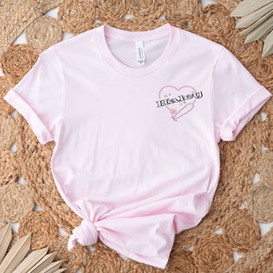 K&ouml;nnte beinhalten: Rosafarbenes T-Shirt mit einem Grafikdruck eines Herzens mit einer Sicherheitsnadel und dem Wort "Haley" darin.