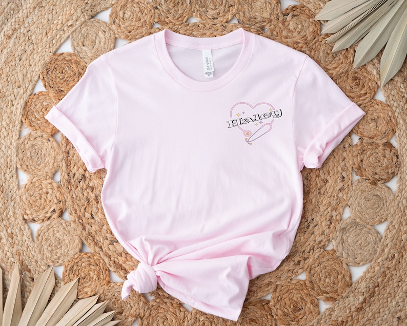 K&ouml;nnte beinhalten: Rosafarbenes T-Shirt mit einem Grafikdruck eines Herzens mit einer Sicherheitsnadel und dem Wort "Haley" darin.