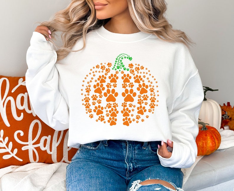 May include: White sweatshirt featuring an orange pumpkin design made of paw prints. The pumpkin stem is green. The sweatshirt is part of a fall-themed collection. An orange pillow with the words "happy fall" is visible.