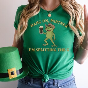 A woman wearing a green t-shirt with a cartoon frog holding a mug and a green hat with a gold square on it.