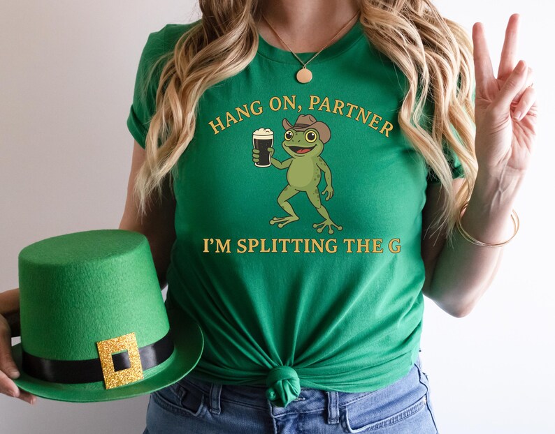 A woman wearing a green t-shirt with a cartoon frog holding a mug and a green hat with a gold square on it.