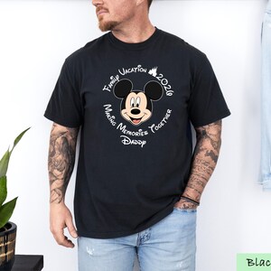 A man wearing a black t-shirt with a Mickey Mouse design and text around it, standing next to a plant.