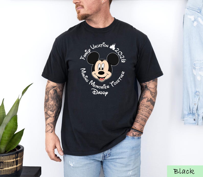 A man wearing a black t-shirt with a Mickey Mouse design and text around it, standing next to a plant.