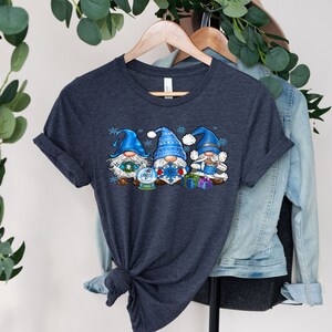May include: A dark heather grey t-shirt with a design of three blue gnomes holding a snow globe, a snowflake, and presents. The gnomes are wearing blue and white hats and have white beards.